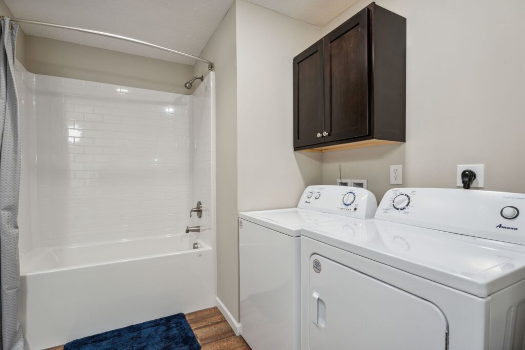 Bathroom with a white bathtub and shower, wall-mounted dark cabinet, and a white washer and dryer set on a wood floor.