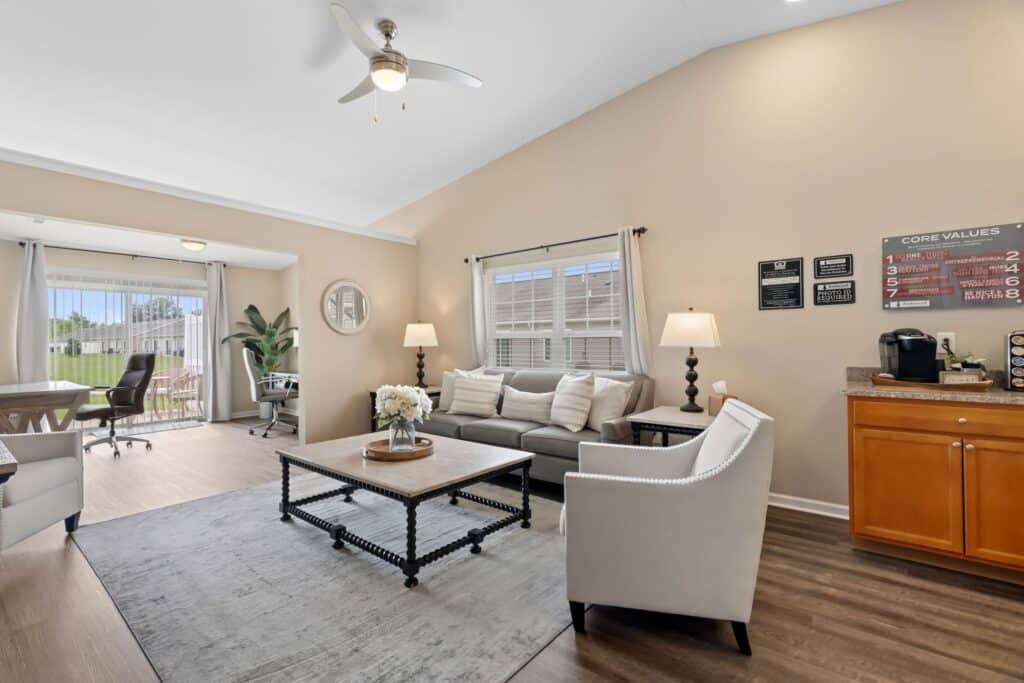 A modern living room with a sofa, armchair, coffee table, and lamps, connected to a dining area with large windows and outdoor views.