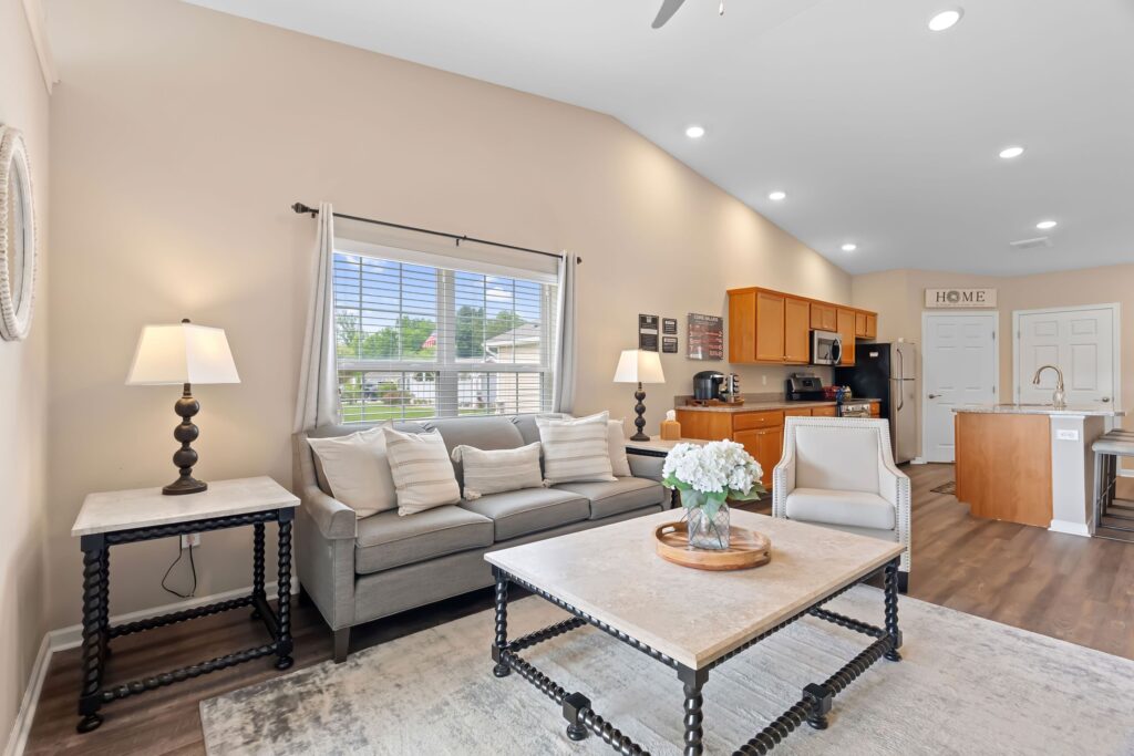 Bright living room with a gray sofa, marble coffee table, lamps, and a window, with an open view to a kitchen featuring wooden cabinets and an island.