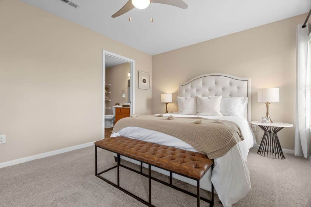 A neatly made bed with a tufted headboard, brown bench, beige walls, two side tables with lamps, and an open door leading to a bathroom.