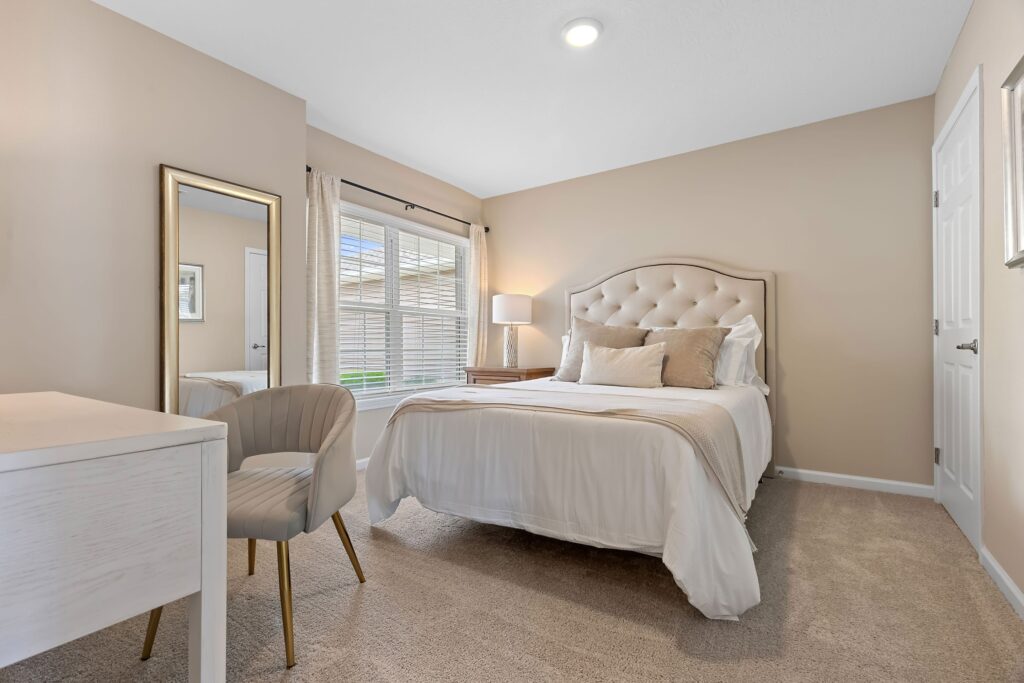 A neatly made bed with beige and white bedding is centered in a carpeted bedroom, with a desk, chair, floor mirror, window, and lamp visible.