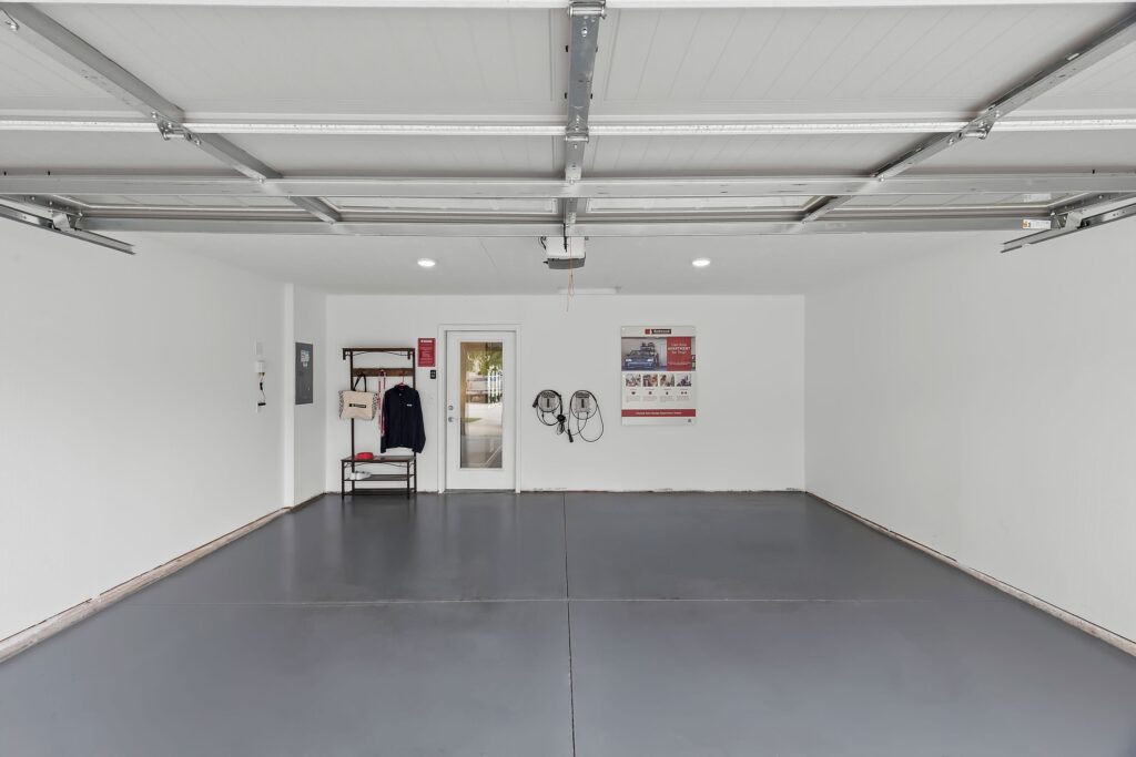 A clean, empty two-car garage with gray floors, white walls, a coat rack with jackets, wall-mounted hoses, a window, and a poster on the back wall.