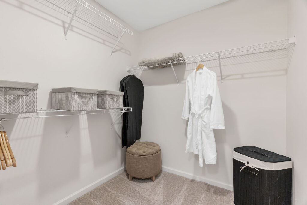 A clean walk-in closet with white wire shelves, storage bins, a hanging white robe, a hamper, hangers, and a beige ottoman.