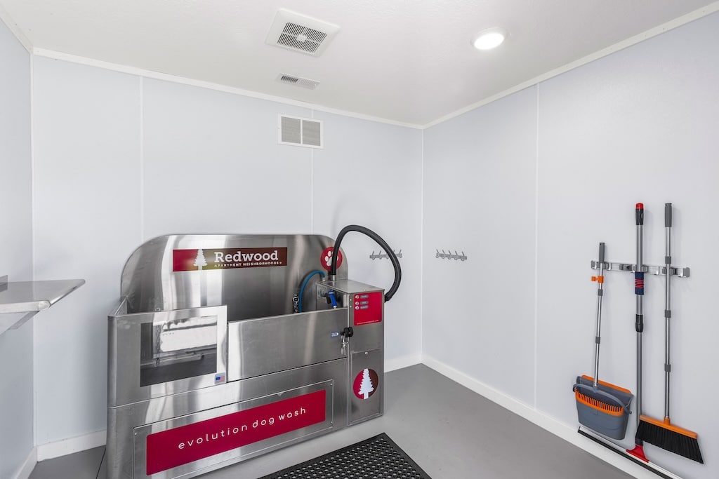 A stainless steel self-serve dog wash station sits against a white wall; cleaning supplies and hooks are mounted nearby in a brightly lit, clean room.