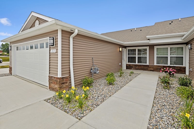 Single-story tan house with attached two-car garage, rock landscaping, yellow flowers, and a concrete walkway leading to the front entrance.