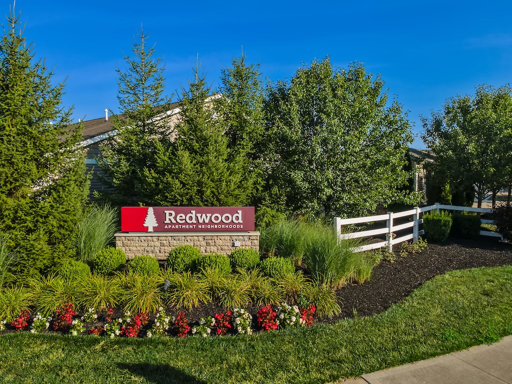 A landscaped entrance with a red sign reading "Redwood Apartment Neighborhoods," surrounded by green shrubs, trees, flowers, and a white fence.