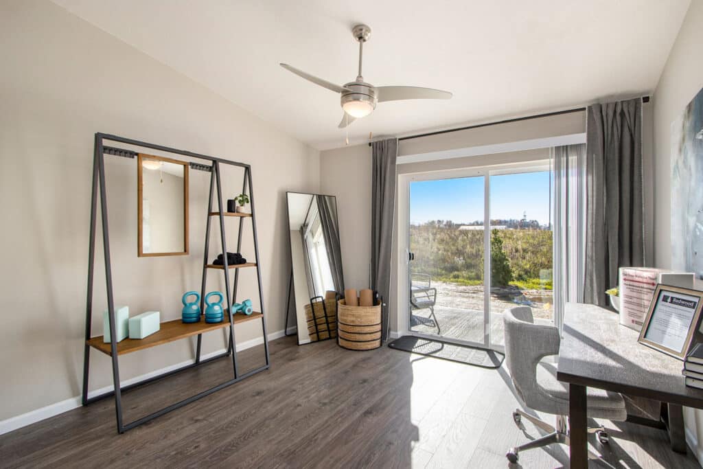 A modern home office with a desk, chair, large mirror, exercise equipment, and sliding glass doors leading to an outdoor area with natural light.