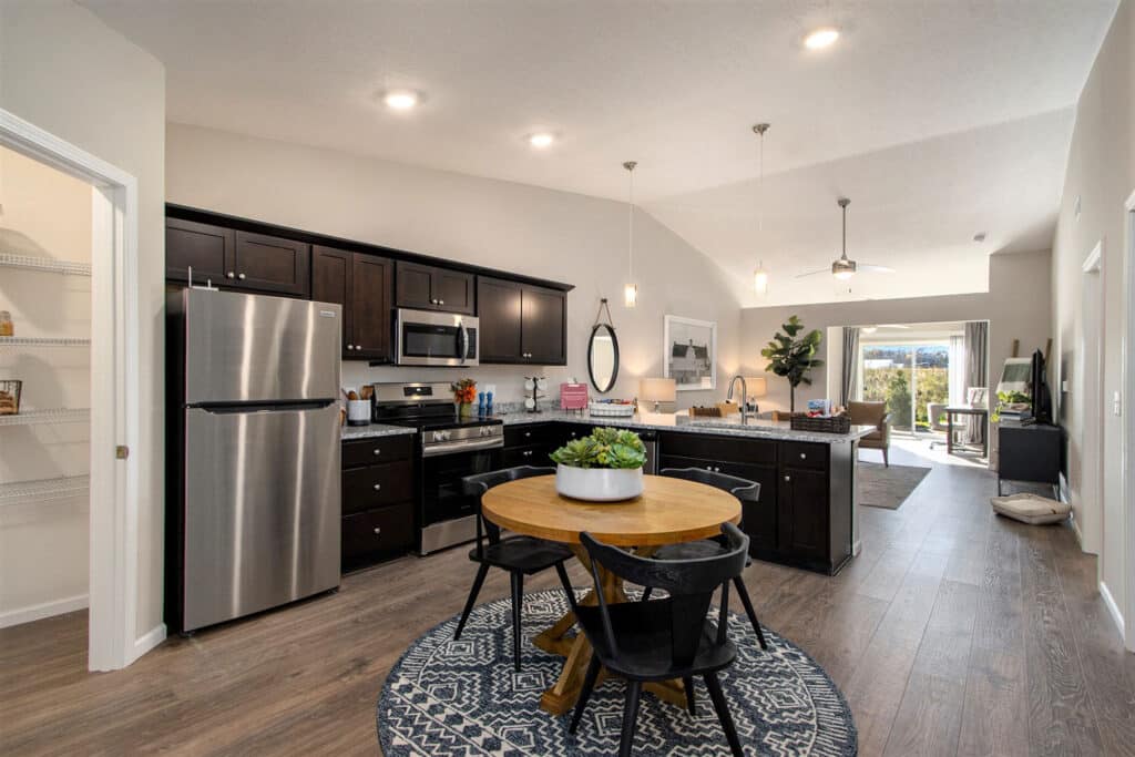 Modern open-concept kitchen and dining area with dark cabinets, stainless steel appliances, round table, and view into a bright living room with a sliding glass door.