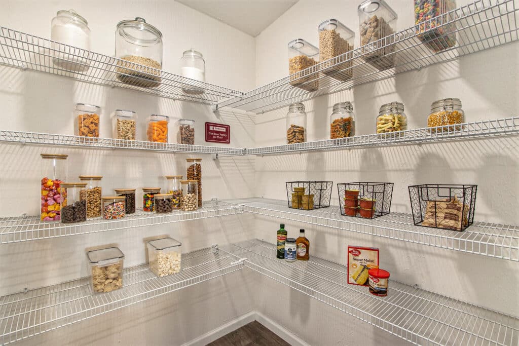 A pantry with wire shelves holding glass jars of snacks, pasta, and grains, baskets with canned goods, and a few bottles and boxes on lower shelves.