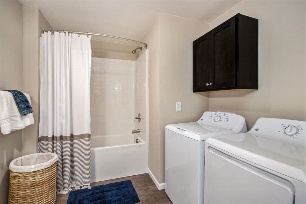 Modern bathroom with a bathtub and shower, white curtain, washing machine, dryer, dark wall cabinet, laundry basket, and towels.