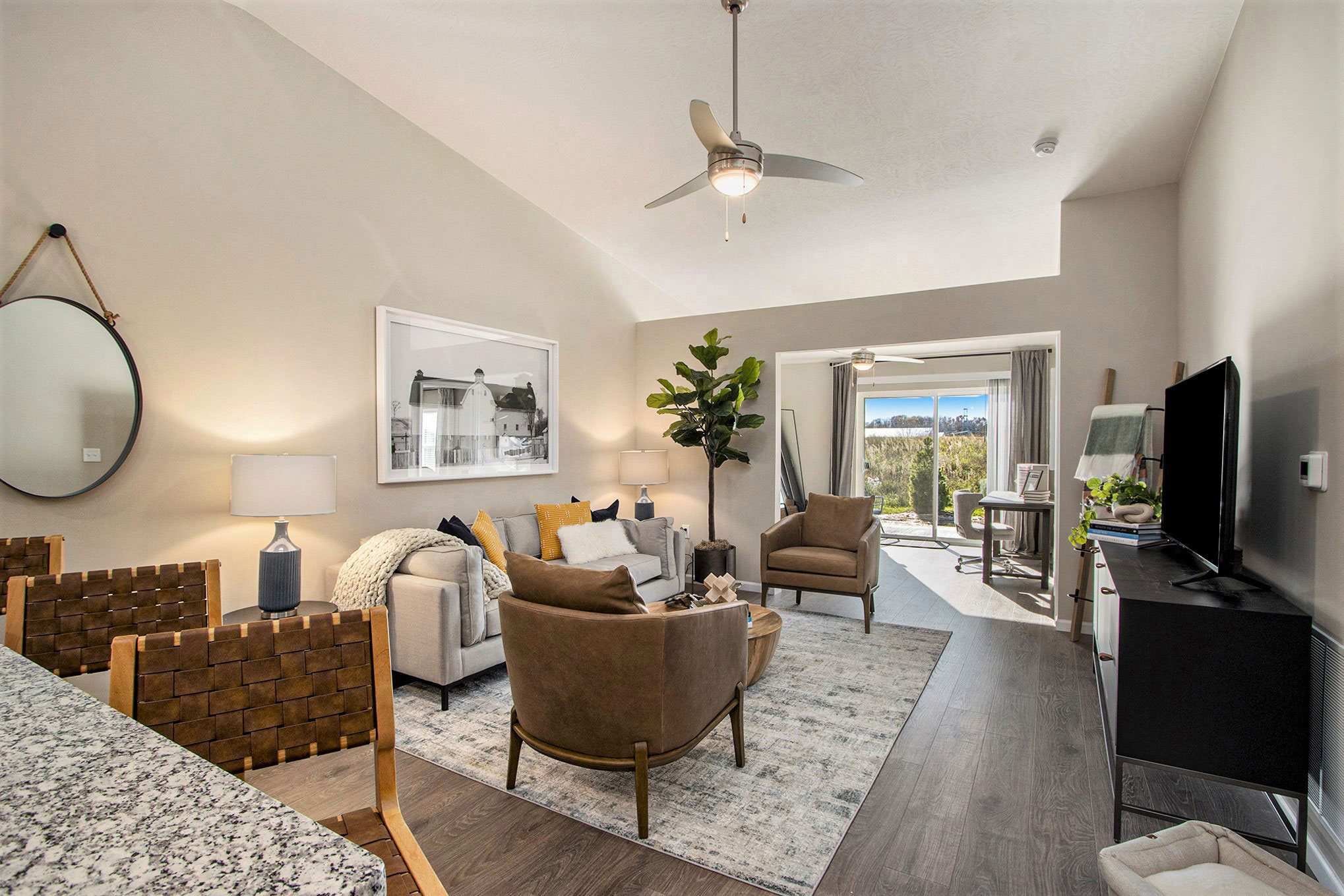 Modern living room with neutral tones features a sofa, two armchairs, wall art, a TV, a ceiling fan, and a large window leading to a sunlit balcony or sitting area.