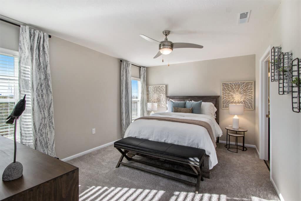 Modern bedroom with a large bed, neutral decor, ceiling fan, two windows with curtains, and a dresser with decorative items.