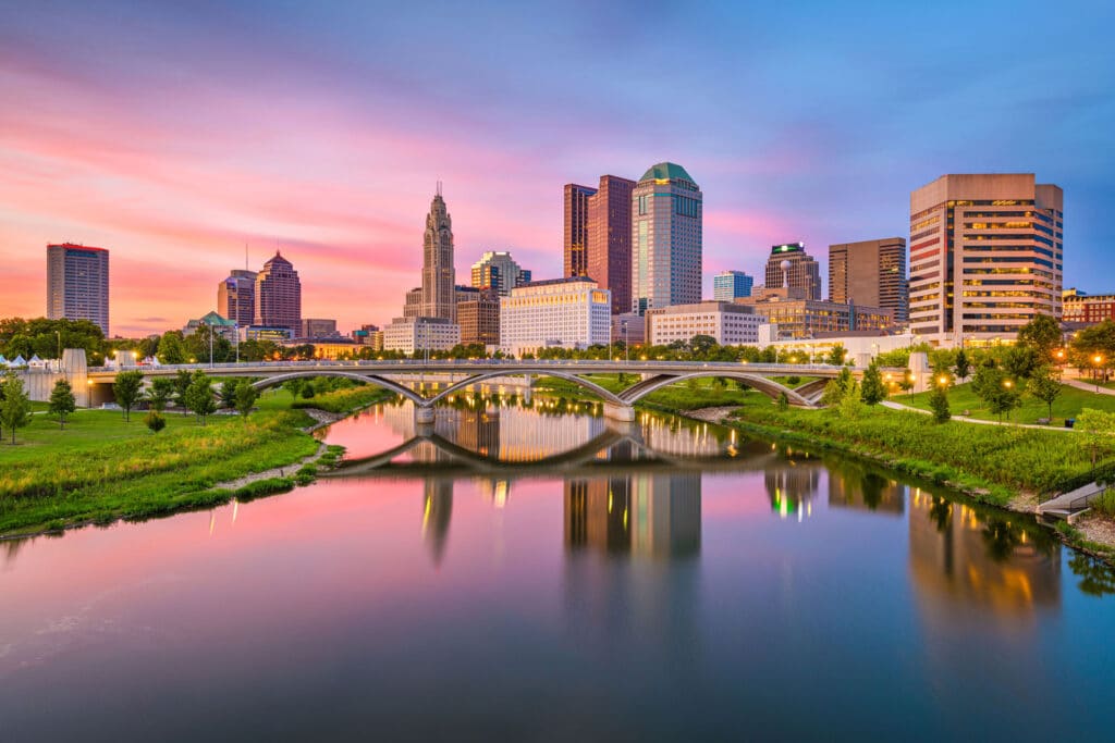 A city skyline at sunset with modern buildings, a river, green spaces, and a bridge reflecting in the water.