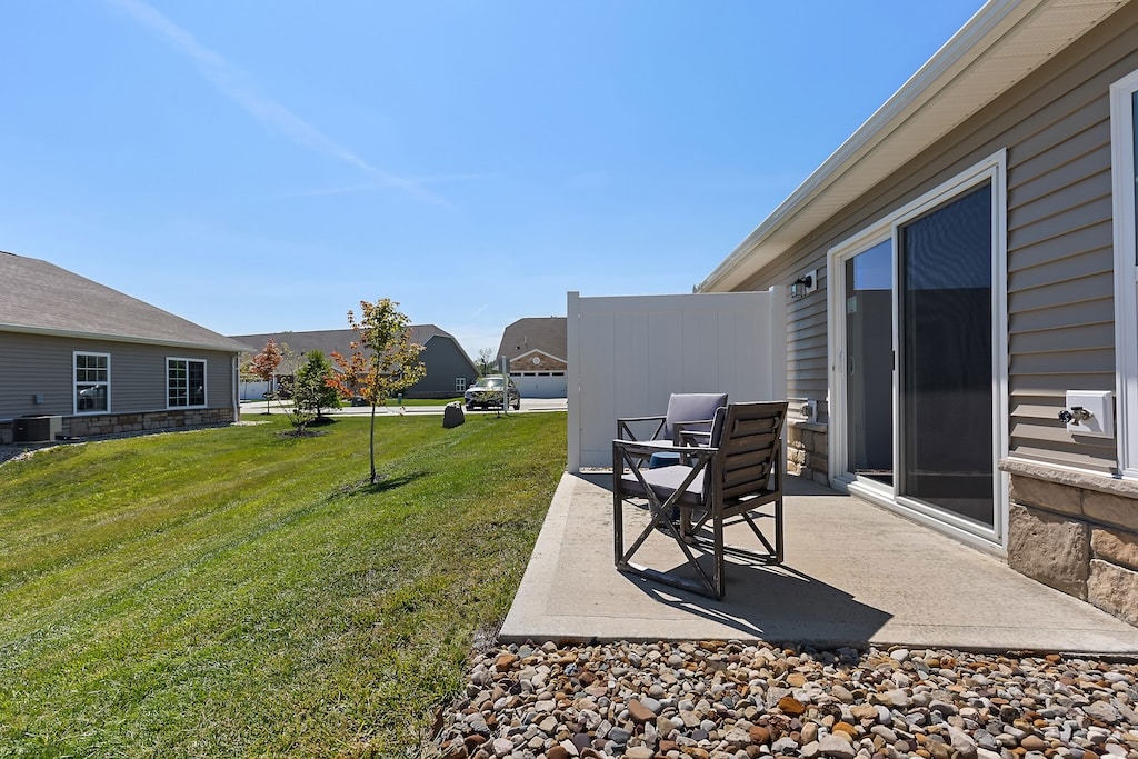 Small concrete patio with two chairs and a side table next to a house, sliding glass door, grassy yard, and a white privacy fence under a clear sky.