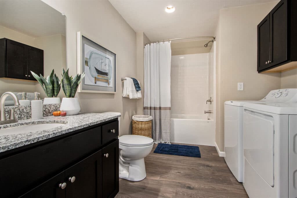 Modern bathroom with dark cabinets, granite countertop, potted plant, toilet, bathtub with shower curtain, washer, dryer, and wood-style flooring.
