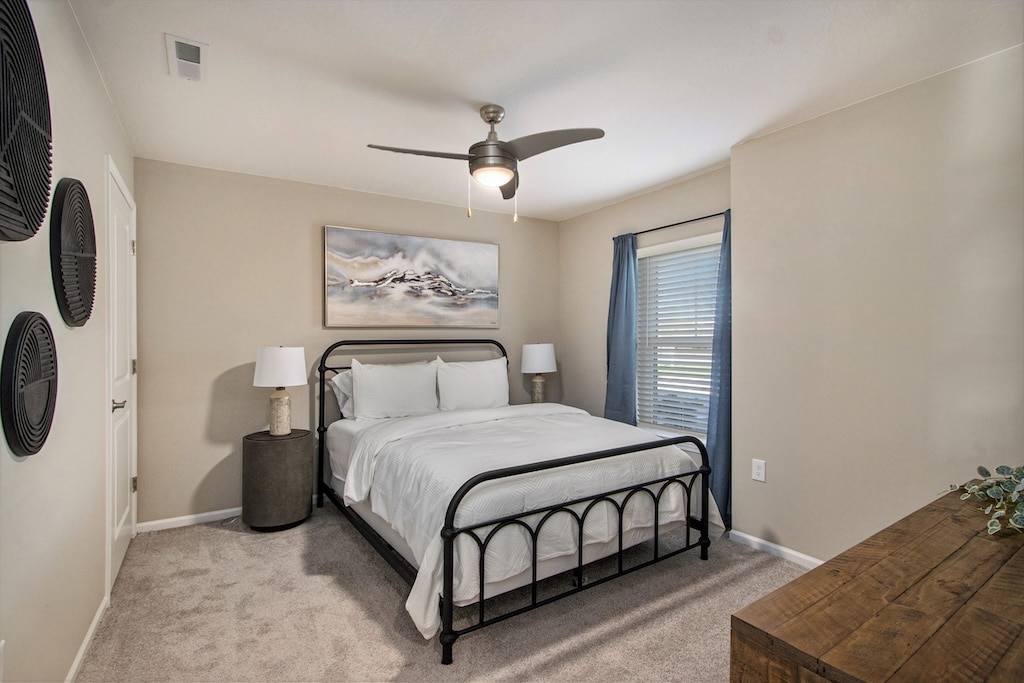 A neatly made bed with white bedding is centered in a simple bedroom with beige walls, a ceiling fan, two nightstands, and a window with blue curtains.