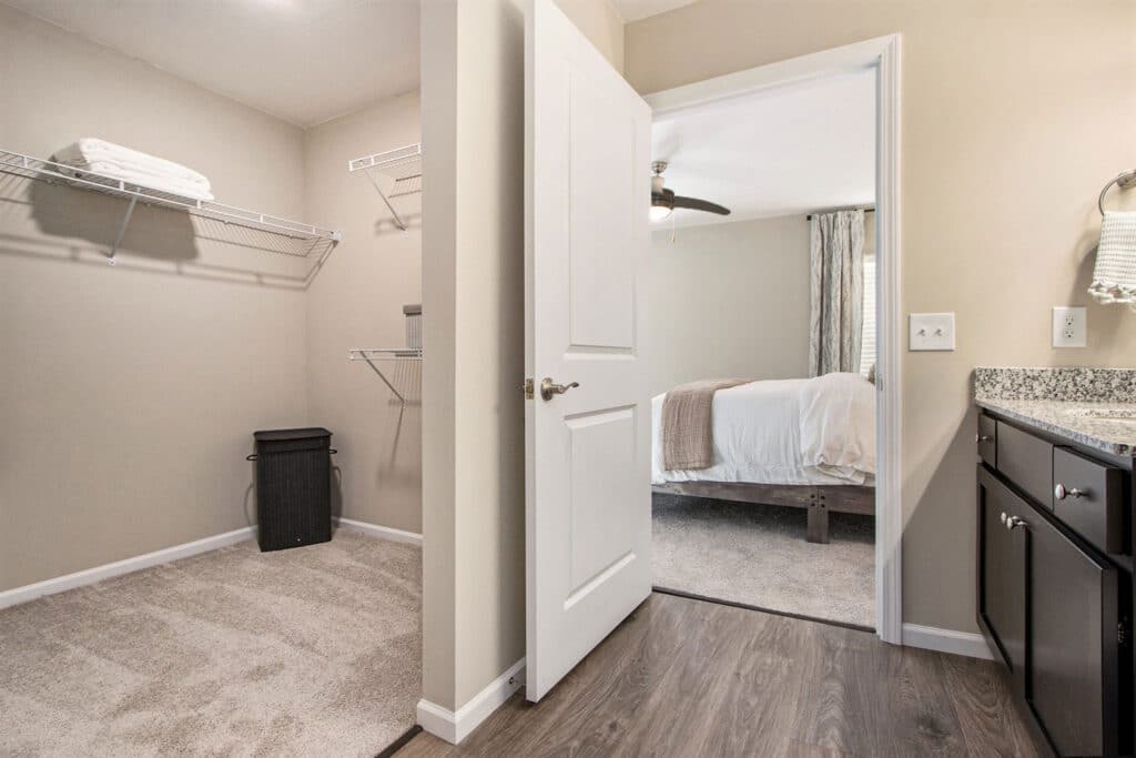 A view from a bathroom shows a walk-in closet with shelves and a laundry basket, and a bedroom with a bed and ceiling fan in the background.