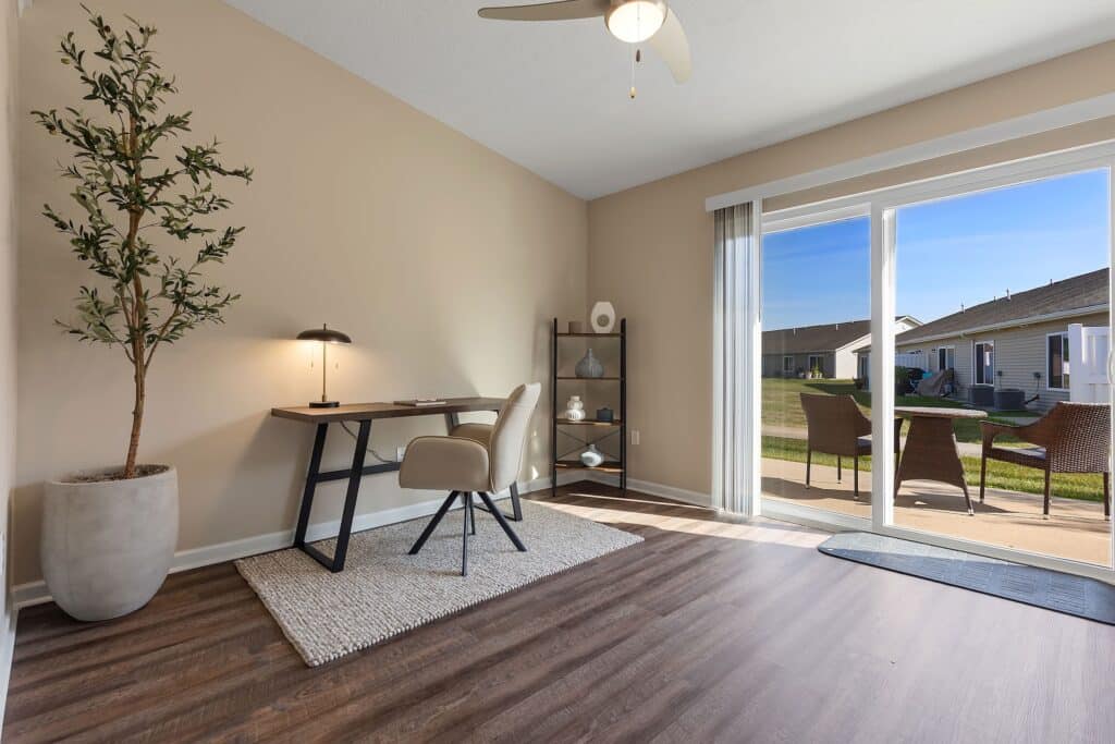 A modern home office with a desk, chair, lamp, and potted plant, featuring a sliding glass door overlooking a patio and yard.