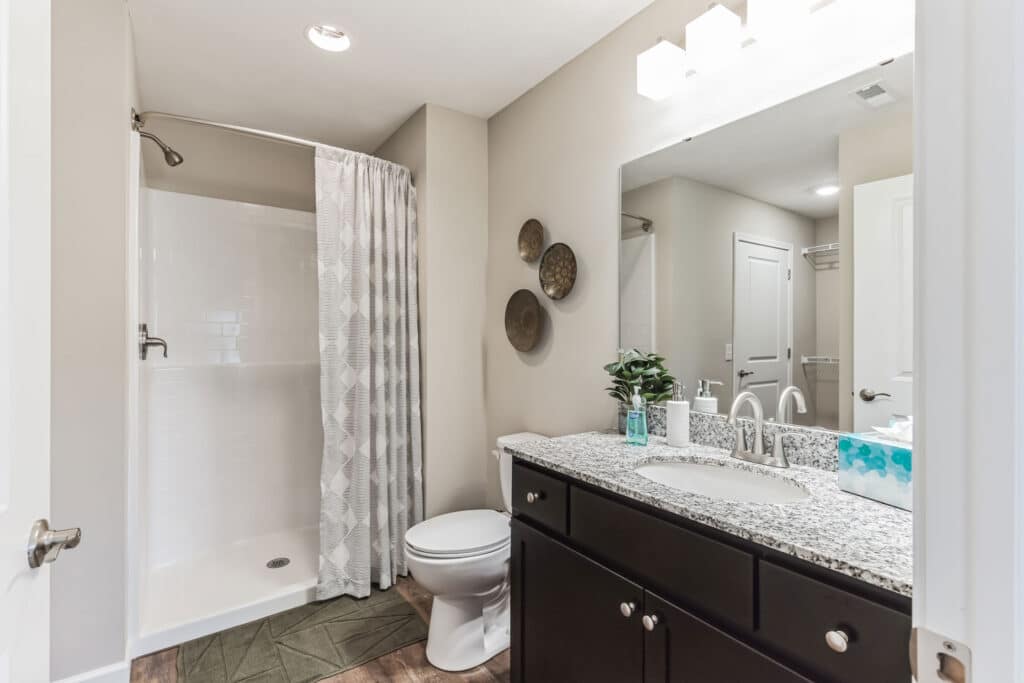 Modern bathroom with a walk-in shower, patterned curtain, toilet, dark vanity with granite countertop, large mirror, and wall-mounted lights.