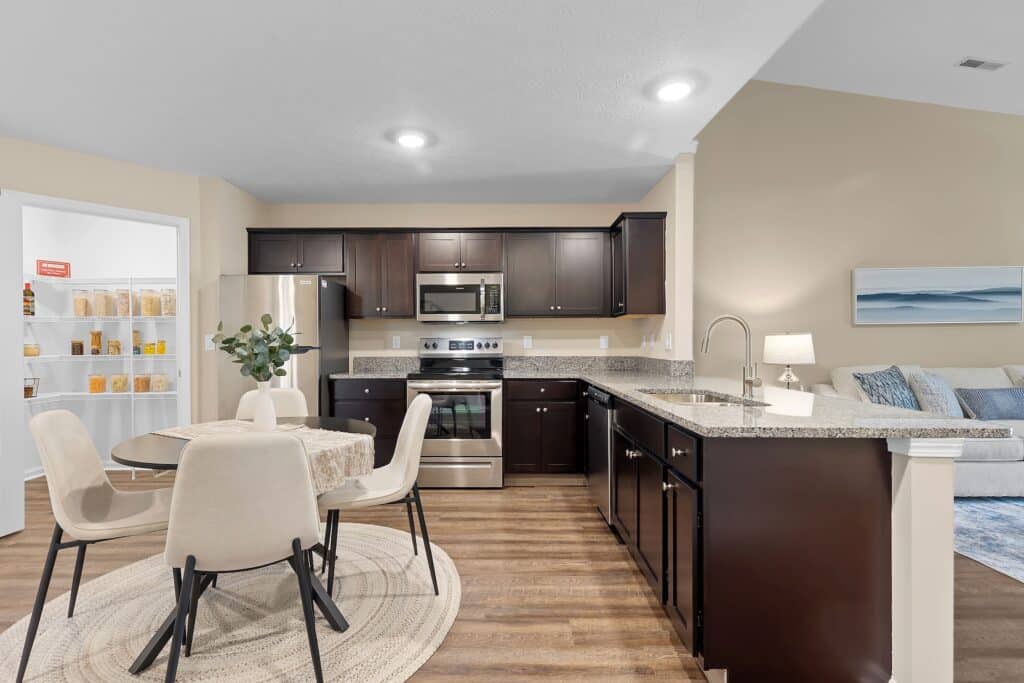 Modern kitchen with dark cabinets, stainless steel appliances, round dining table with four chairs, and a visible pantry. Living area with couch and painting in the background.