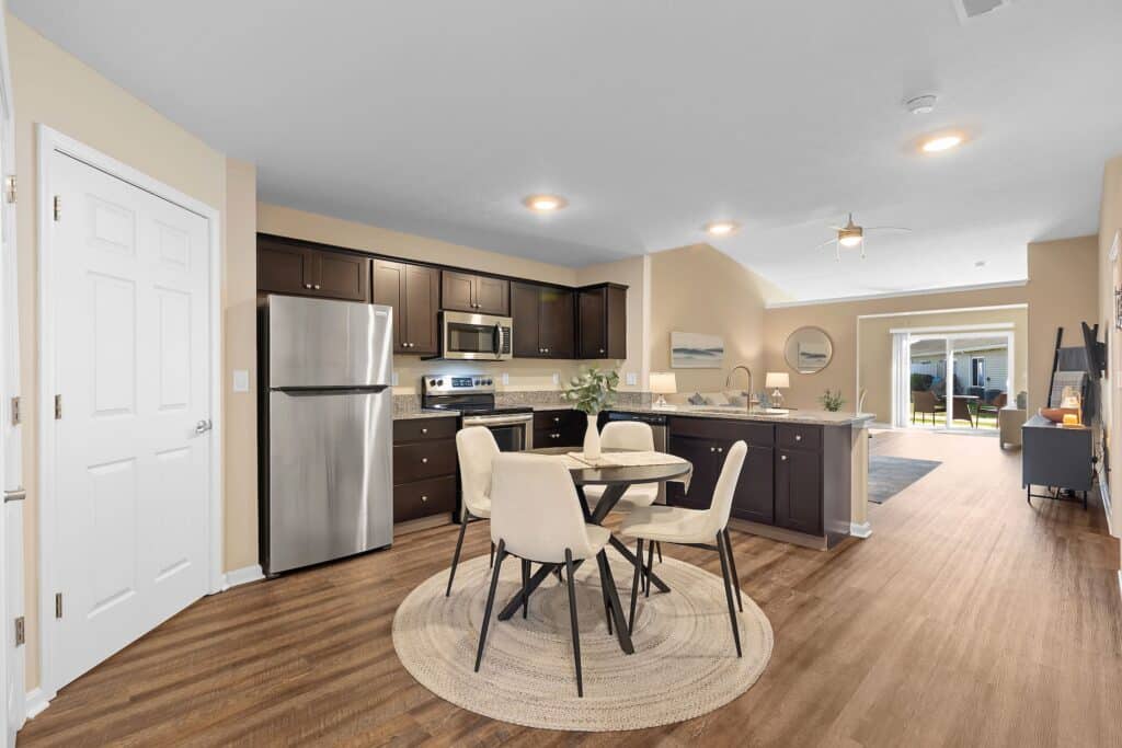 Modern kitchen and dining area with stainless steel appliances, dark cabinets, round table with four chairs, and wood flooring, opening to a living area and patio.