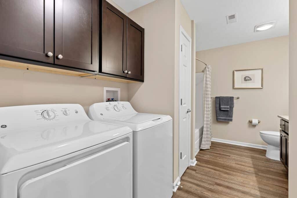 A laundry area with a washing machine and dryer next to a bathroom with a tub, toilet, towel rack, and wall cabinets, all on wood flooring.
