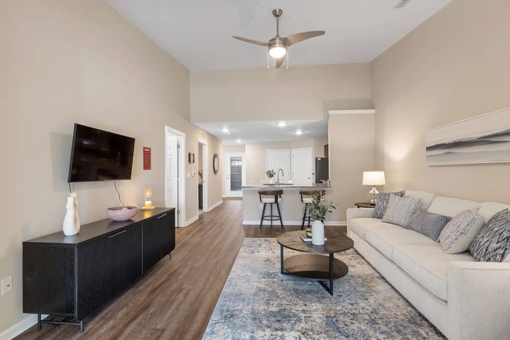 Modern living room with a beige sofa, round coffee table, wall-mounted TV, and open view into a kitchen with bar seating under recessed lights.