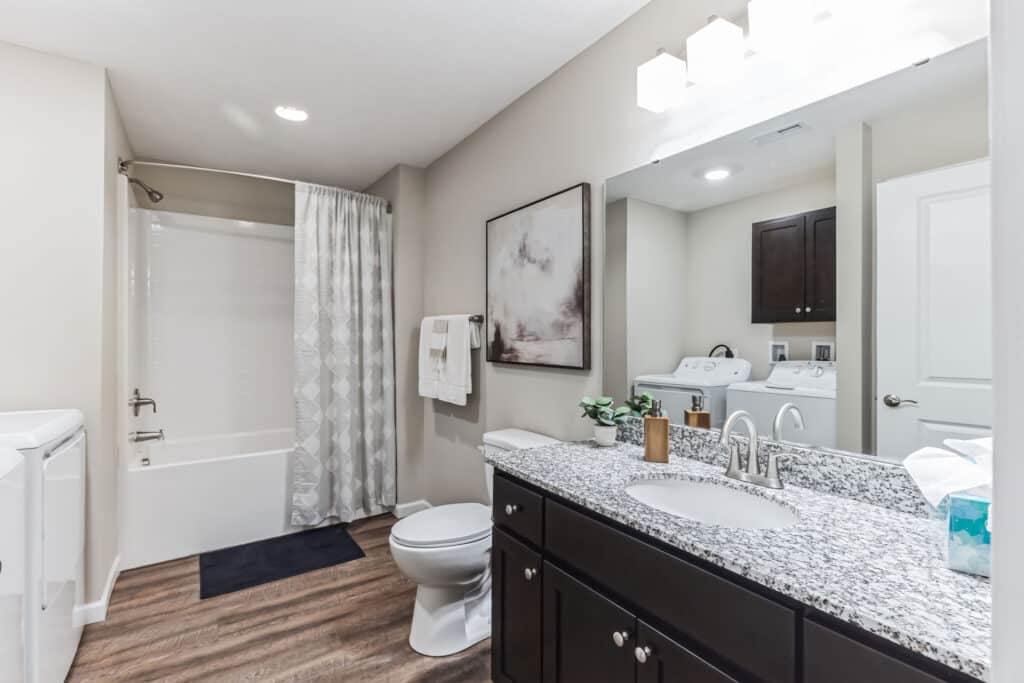 Modern bathroom with a granite countertop, double sinks, a toilet, bathtub with shower curtain, wall art, and a washer and dryer in the corner.