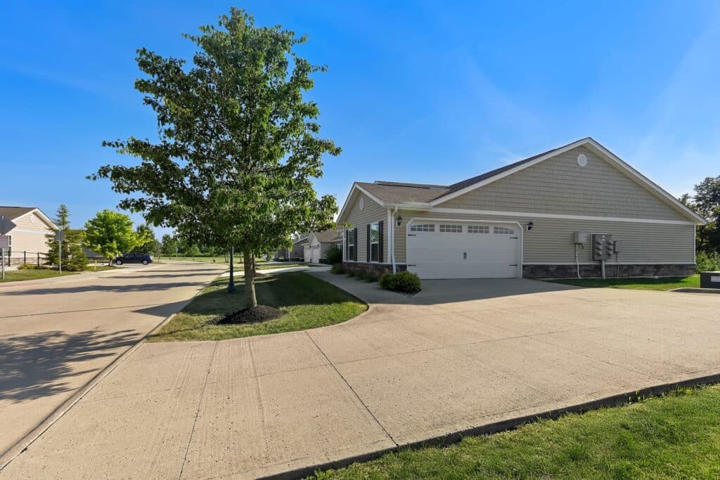 Single-story house with a double garage, tan siding, and a large driveway, located on a suburban street with a green lawn and a tree in front.