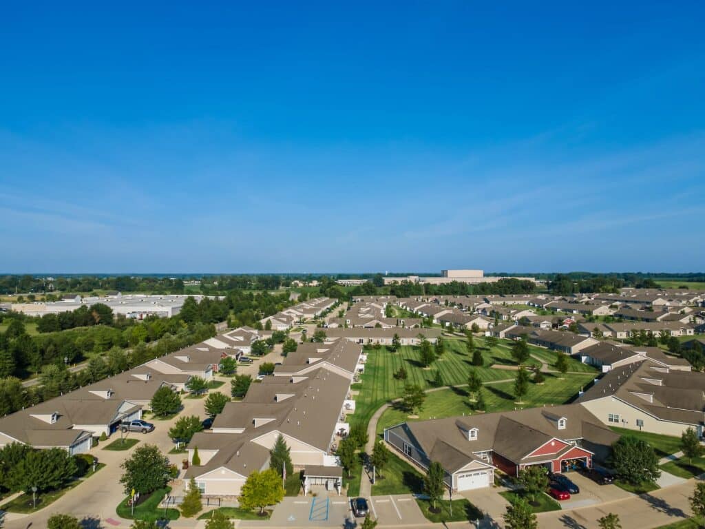 Aerial view of a suburban residential neighborhood with rows of houses, green lawns, trees, and clear blue sky.
