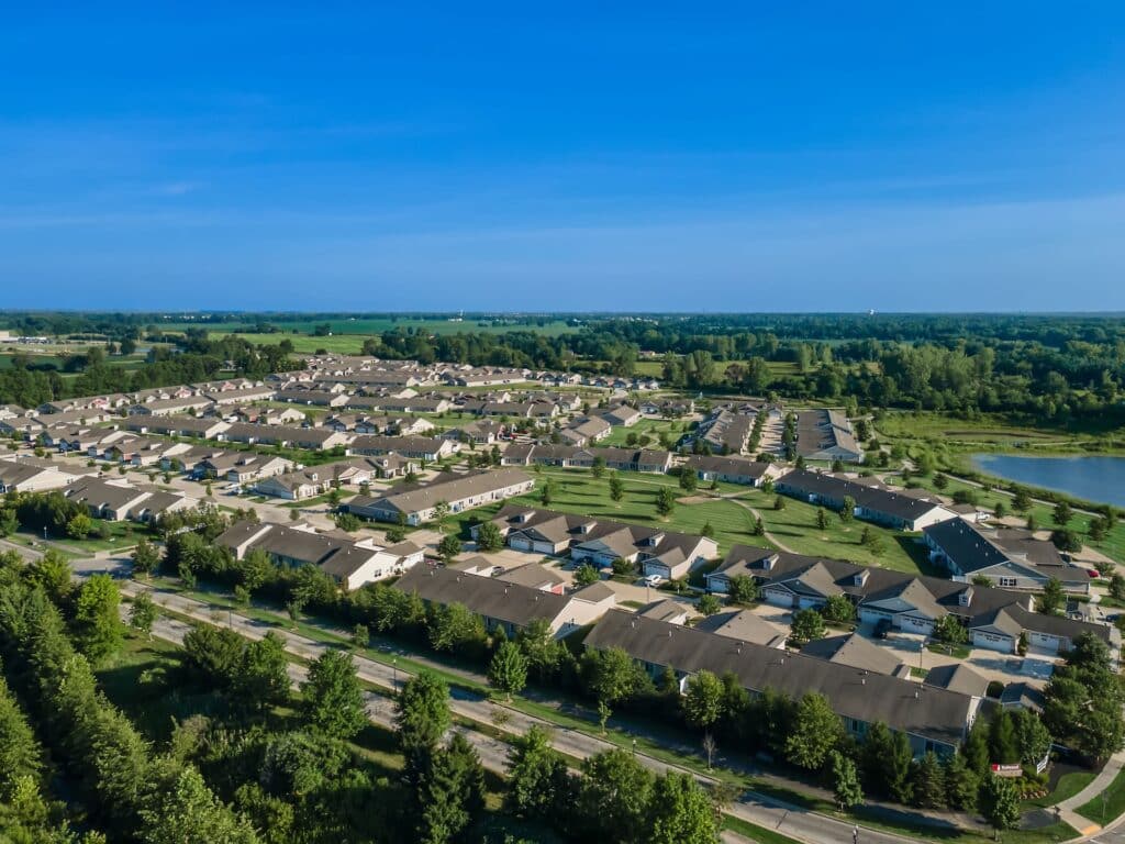 Aerial view of a suburban neighborhood with rows of houses, green lawns, trees, a road in the foreground, and open fields in the distance under a clear blue sky.