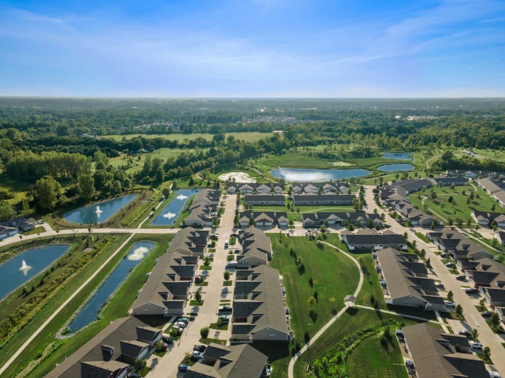 Aerial view of a suburban neighborhood with rows of houses, green lawns, ponds with fountains, and surrounding trees under a clear blue sky.