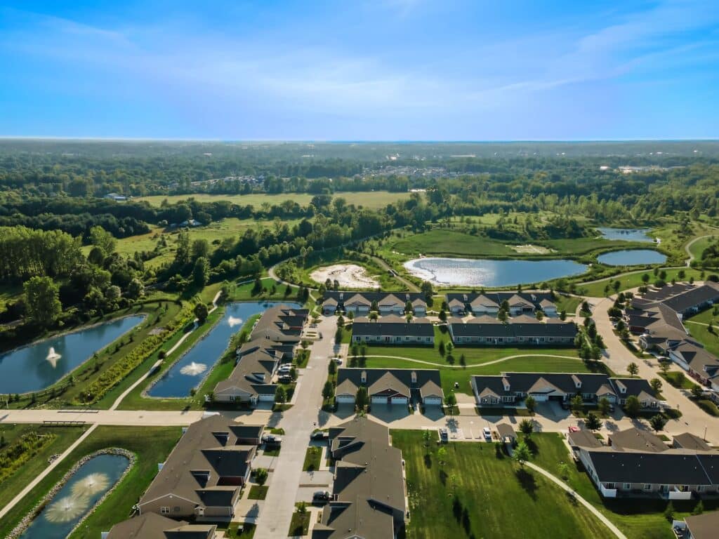 Aerial view of a suburban neighborhood with rows of houses, streets, small lakes, fountains, and green spaces under a clear blue sky.
