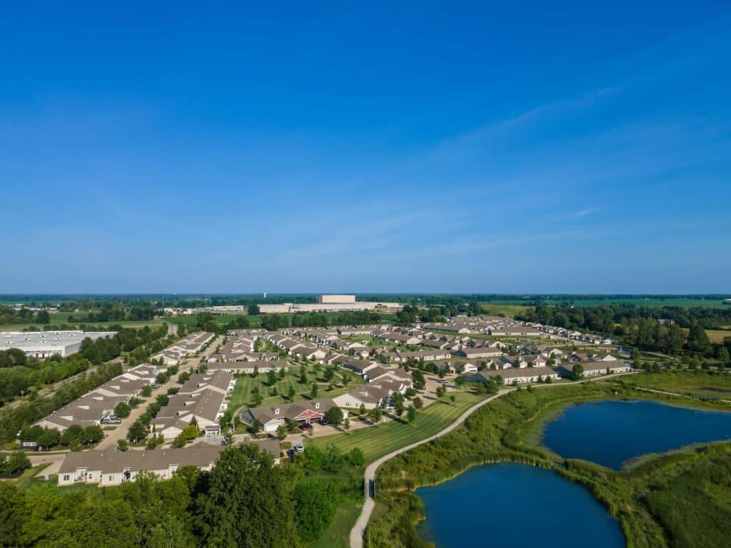 Aerial view of a suburban residential neighborhood with rows of houses, green lawns, and ponds under a clear blue sky.