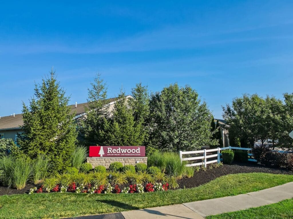 A landscaped entrance with a red "Redwood Apartment Neighborhoods" sign, surrounded by bushes, flowers, trees, and a white fence under a clear blue sky.