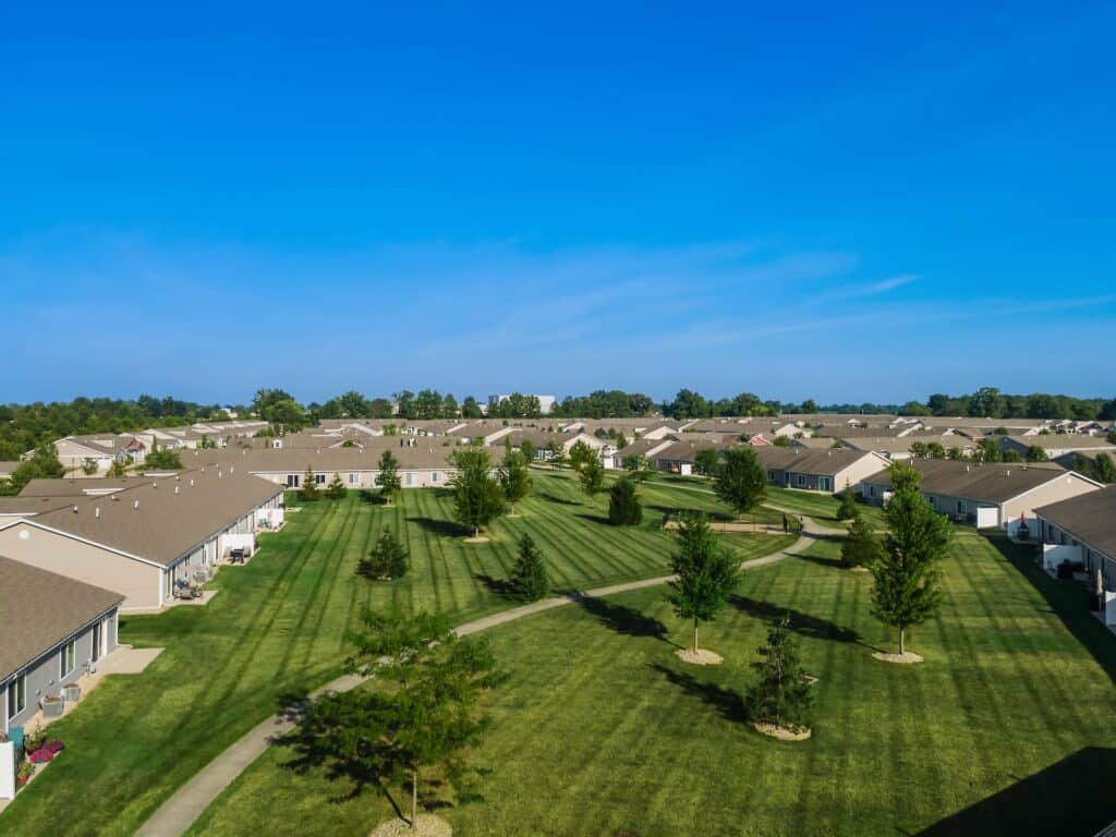 Aerial view of a residential neighborhood with rows of single-story houses, neatly mowed lawns, young trees, and clear blue sky.