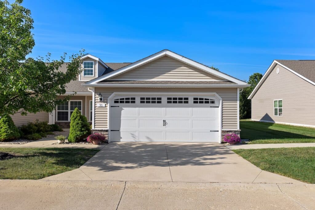 Single-story suburban house with beige siding, a white double garage door, small landscaping, and a clear blue sky overhead.