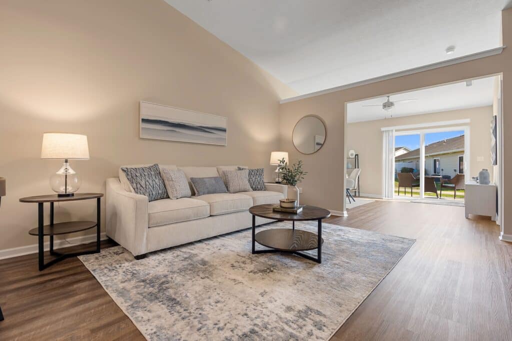 A modern living room with a beige sofa, two round coffee tables, a table lamp, wall art, and a rug. Large glass doors open to a patio and let in natural light.
