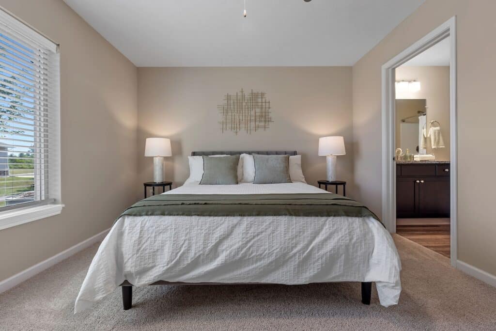 A neatly made bed with white linens and green accents sits between two lamps in a beige bedroom, with a bathroom visible through an open door on the right.