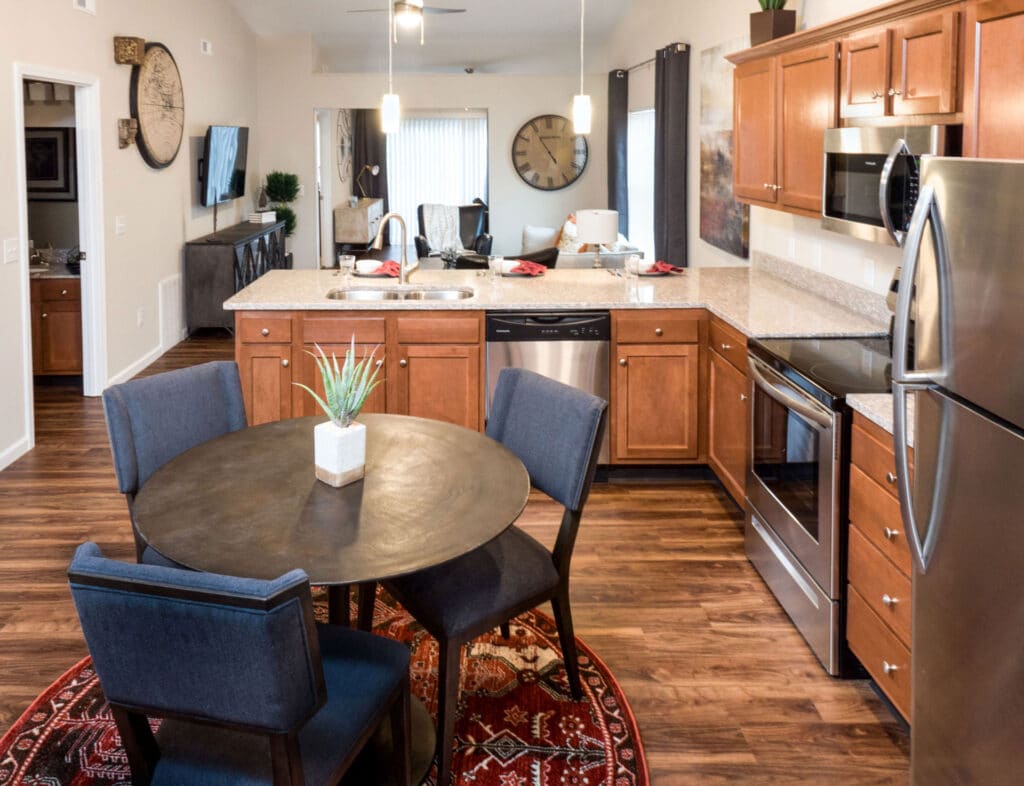 Modern kitchen and dining area with stainless steel appliances, wooden cabinets, a round dining table with four blue chairs, and a decorative plant centerpiece on a patterned rug.