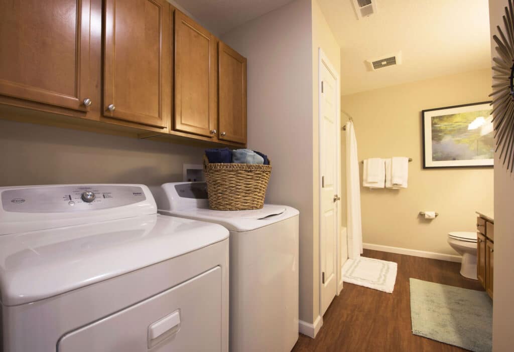 Laundry room with a washer and dryer, wooden cabinets above, a basket on the dryer, and a bathroom visible in the background with towels, a mat, and wall art.