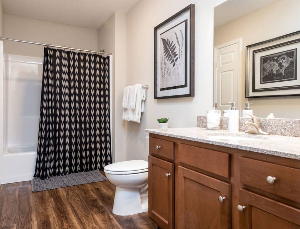 Modern bathroom with wood flooring, brown cabinets, granite countertop, white toilet, black-and-white shower curtain, and framed artwork on the walls.