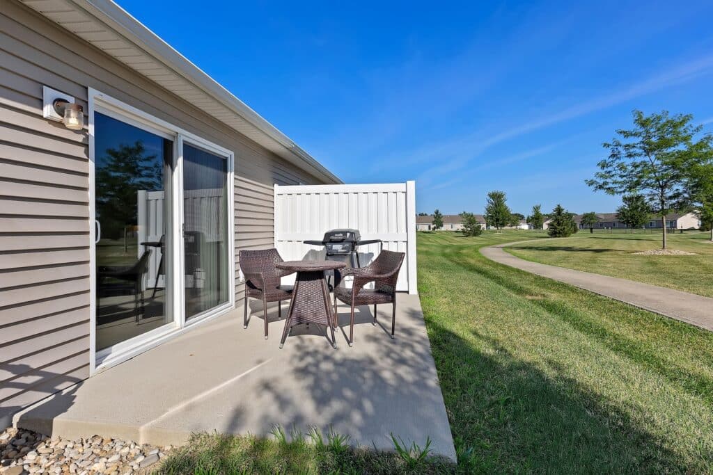 A small concrete patio with a round table, four wicker chairs, and a barbecue grill, next to a sliding glass door and a white privacy fence, overlooking a grassy yard and sidewalk.