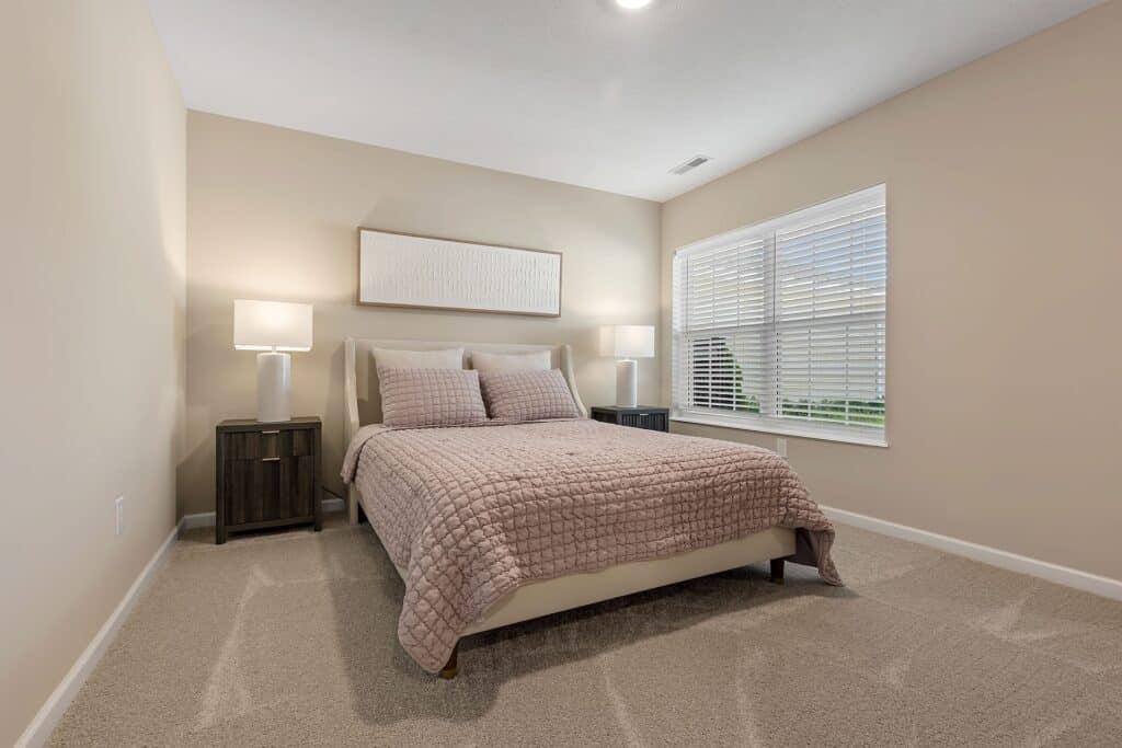 A neatly made bed with pink bedding is centered between two dark nightstands with white lamps in a beige-walled bedroom with carpet and a large window.