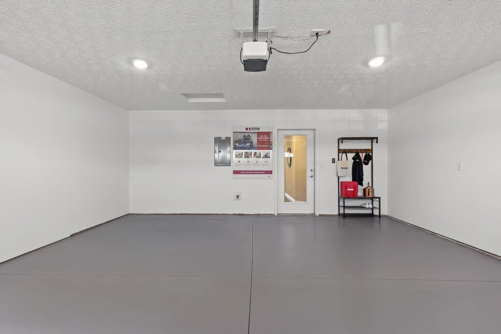 A clean, empty garage with white walls, gray floor, wall-mounted control panels, a door, a window, a poster, and a small shelving unit with household items.