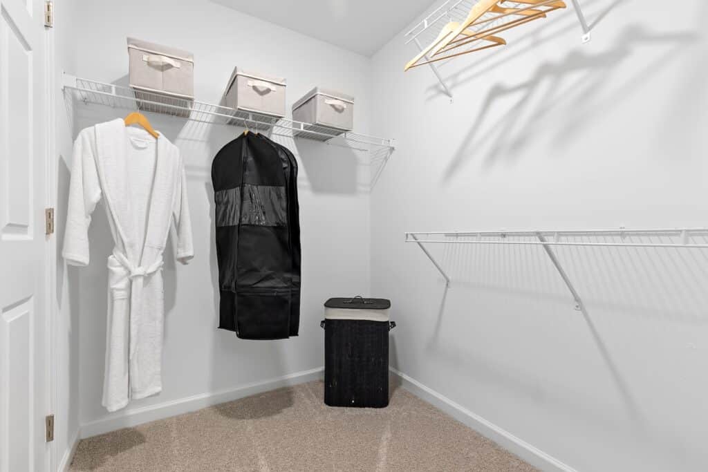 A walk-in closet with a white robe, a black garment bag, beige storage boxes on a shelf, wooden hangers, and a black laundry basket on a carpeted floor.