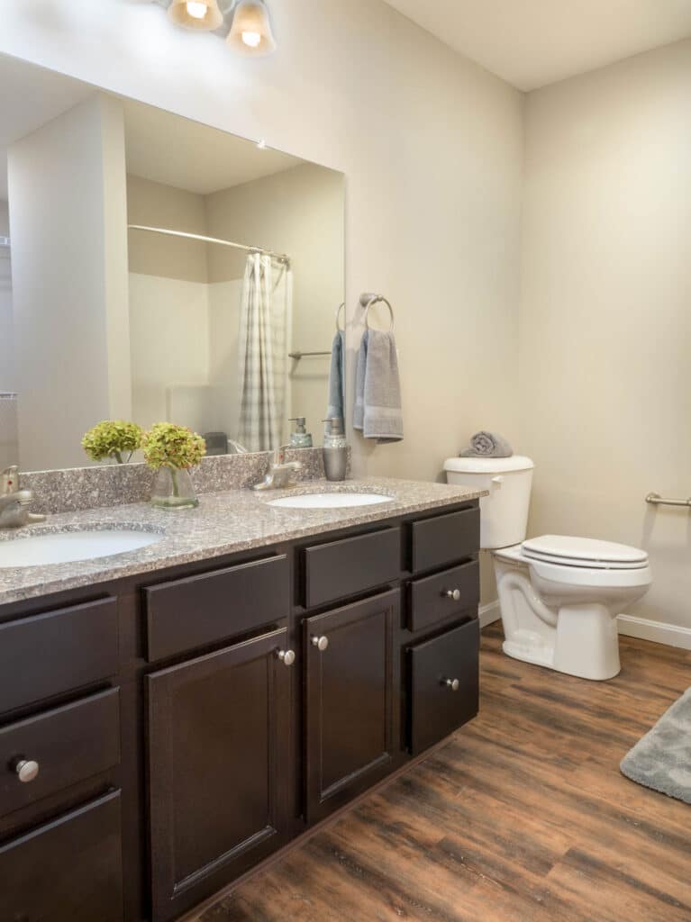 Modern bathroom with double-sink dark wood vanity, granite countertop, flowers, toilet, towel rack, and wood flooring. Neutral walls and a large mirror are visible.