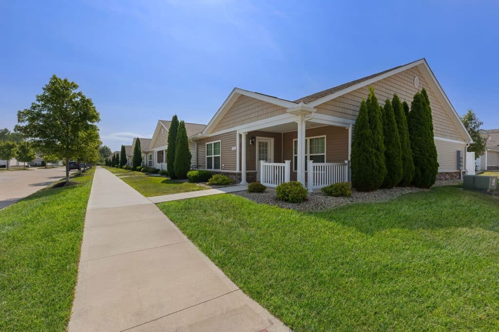 Row of single-story suburban houses with front porches, manicured lawns, and a sidewalk under a clear blue sky.