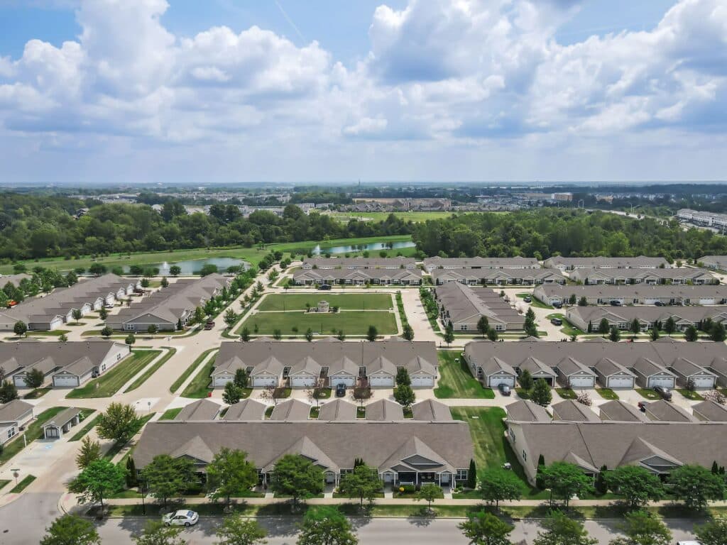 Aerial view of a suburban residential neighborhood with rows of houses, green lawns, trees, and a central open space under a partly cloudy sky.