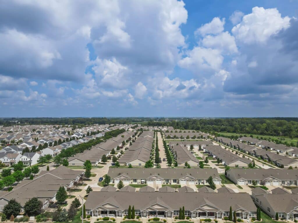 Aerial view of a suburban neighborhood with rows of identical houses, neatly aligned streets, and a backdrop of trees under a partly cloudy sky.